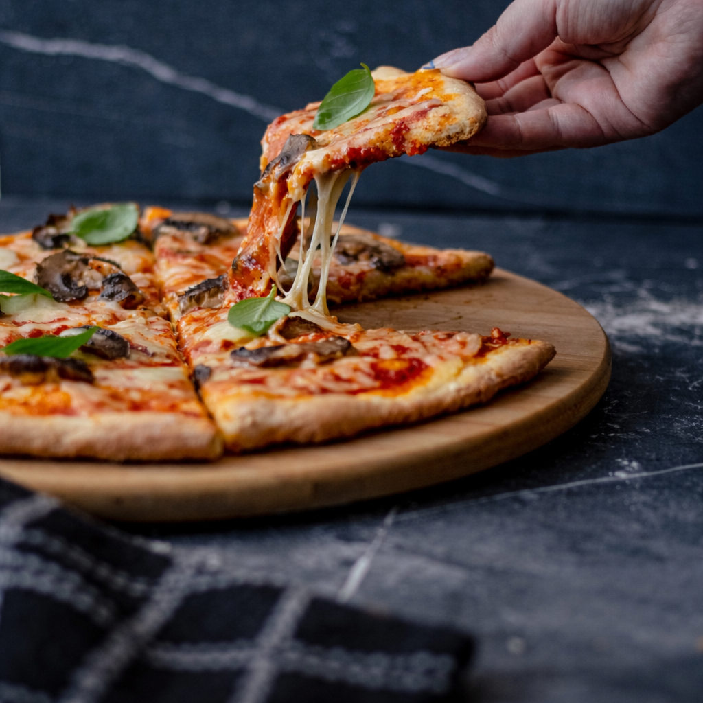 Pizza with a slice being lifted, showing melted almond mozzarella, on a wooden board against a dark background.